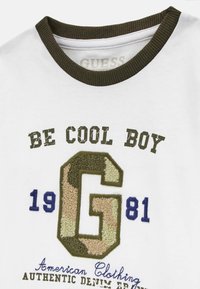 T-shirt en coton blanc avec col côtelé vert olive, comportant un texte brodé et un design "G" en camouflage. Détails supplémentaires en bleu et noir.