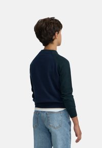 Suéter azul marino con mangas raglán, puños y dobladillo de canalé, hecho de tela ligera, usado sobre unos jeans de mezclilla, visto desde la espalda.