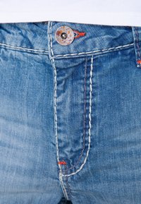 Jeans de mezclilla azul con lavado claro, que cuentan con un cierre de botón, costuras visibles en blanco y azul oscuro, y acentos bordados en rojo.