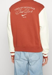 Nike sportströja med en rostfärgad kropp, krämfärgade ärmar och "Just Do It"-emblem på baksidan, samt ribbade detaljer.