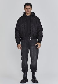 Man in een zwart gewatteerd jack over een zwarte hoodie, met donker gewassen jogger jeans en zwarte chunky boots, staand tegen een lichtgrijze achtergrond.