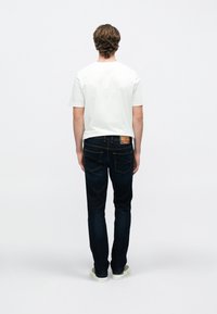 Bela majica s kratkimi rokavi, kombinirana s temno modrimi jeans hlačami. Jeans hlače imajo kontrastni rumeni šiv in barvno znamko na zadnji strani.