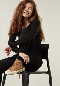 Maglietta nera a maniche lunghe e leggings neri, abbinati a sneaker alte marrone chiaro. Modella seduta su una sedia nera, con uno sfondo neutro.