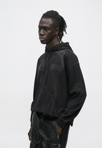 Jauns melnādainais vīrietis ar drediem, ģērbies melnā Diesel džemperī un melnās ādas kravāžas biksēs, stāv ar rokām kabatās uz baltas fona.