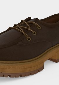 Gros plan d'une chaussure en daim marron avec des lacets ronds, des œillets métalliques et une semelle épaisse en caoutchouc texturé avec motif de bande de roulement.