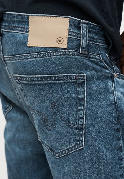 Κοντινό πλάνο πίσω τσέπης μπλε τζιν με ραμμένο σχέδιο κυμάτωσης και ταμπέλα από ταμπά δέρμα με την ένδειξη «AG» στη μέση.