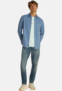 Camicia celeste a maniche lunghe indossata sopra una t-shirt verde menta, abbinata a jeans di lavaggio medio e sneaker beige. Presenta una tasca sul petto.