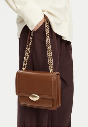 Braune Leder-Schultertasche mit goldener Kettenriemen und ovalem goldfarbenem Verschluss, getragen von einer Person mit beigem Oberteil und dunkelbrauner Hose.