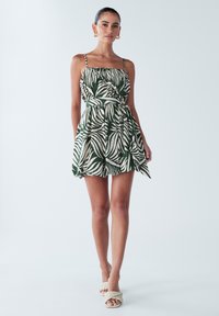 Bezroku kleita ar zaļu un baltu palmas lapu rakstu, pieguļošs korsete, saplacināta vidukļa daļa un izplūdināts svārks. Redzams siksnu dizains un auduma tekstūra.