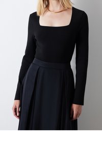 Top nero a maniche lunghe con scollatura quadrata abbinato a una gonna nera plissettata. Il tessuto sembra liscio e uniforme, senza motivi.