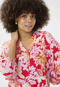 Raudona gėlėta bluzė su laisvu siluetu, turinti V formos kaklą, mygtukų užsegimą ir pūstas rankoves. Gėlės rožinėmis, oranžinėmis ir grietinėlės spalvomis.