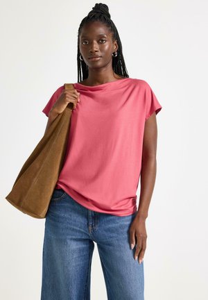 Kvinde med fletninger iført en lyserød kortærmet top, blå jeans, store øreringe og bærende en stor brun skuldertaske.