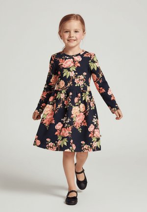 Granatowa sukienka w kwiaty z długimi rękawami, zdobiona różowymi i zielonymi różami, dopasowanym gorsetem i rozkloszowaną spódnicą; zestawiona z czarnymi butami.