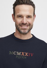 Sudadera navy con cuello redondo, texto bordado "MCMXXIV FQ1924" en verde, naranja y rojo. Tejido suave y sutil textura.