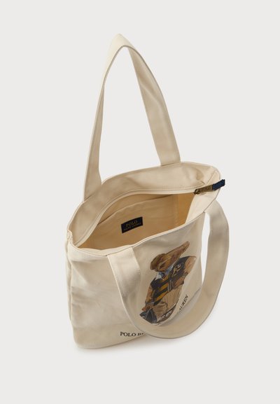 Sac fourre-tout en toile beige avec deux anses, ouverture supérieure et ours en peluche imprimé portant une tenue polo sur le devant.