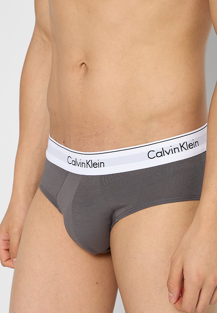 Torso masculino vestido con bóxers grises de Calvin Klein con cintura blanca y logo negro, de pie contra un fondo liso.