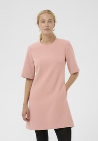 Kurzärmliges rosa Kleid aus strukturiertem Stoff mit rundem Ausschnitt und Seitentaschen, kombiniert mit schwarzen Leggings.