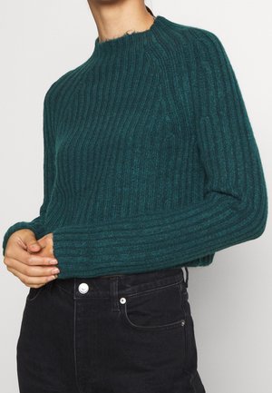 Femme portant un pull en maille côtelée vert foncé avec un col haut et des manches longues, associé à un jean noir taille haute.