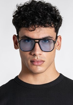 Ung mand med krøllet mørkt hår iført store blåtonede briller, sort ribstrikket trøje og lille ørering mod en ensfarvet lys baggrund.