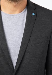 Ciemnoszara marynarka z klapkami i jedną kieszenią, nałożona na jasnoszary t-shirt. Posiada mały niebieski akcent w postaci guzika.