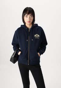 Námornícka mikina na zips so zlatým logom a detailami, ktorej súčasťou je klokanková kapsa a kapucňa so šnúrkou. V kombinácii s čiernymi nohavicami a čiernou taškou.