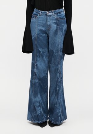 Jean large taille haute bleu avec motifs effet délavé foncé, coutures avant, associé à un haut noir à manches longues avec poignets évasés et des chaussures noires pointues.