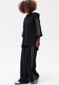 Czarna bluza z szerokimi rękawami, z haftowanymi paskami. Połączona z luźnymi czarnymi spodniami, obie z miękkiego materiału. Widać wygodne obuwie.