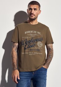 Olivově zelené bavlněné tričko s krátkým rukávem, s modrým grafickým logem/designem a textem v různých písmách. Nalezeno v kombinaci s modrými džíny.