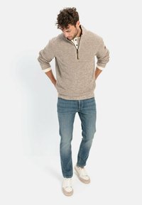 Lichtbruin gebreid sweater met een ritssluiting hals, crème accenten bij de kraag en mouwen, gecombineerd met blauwe jeans en beige sneakers.