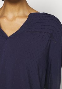 Blouse marine avec un décolleté en V, présentant un tissu texturé avec de petits motifs brodés. Des fronces plissées aux épaules ajoutent des détails.