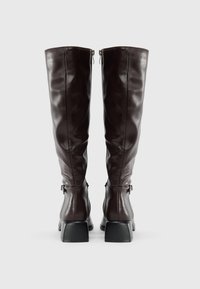 Bottes en cuir marron montantes, avec une finition brillante, des talons carrés et des fermetures éclair latérales, mettant en valeur une texture lisse et un design soigné.