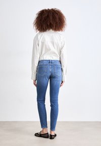 Un par de jeans de mezclilla azul claro con corte ajustado, que cuentan con bolsillos traseros y una cintura alta, combinados con una camisa de rayas de manga larga y zapatos planos negros.