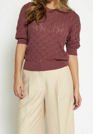 Kvinde iført en tekstureret lilla kortærmet sweater og beige højtsiddende bukser med vide ben mod en hvid baggrund.