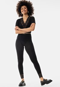 Body noir avec décolletage en dentelle, associé à un legging noir avec des accents en dentelle au niveau des chevilles, et des chaussures noires à enfiler avec des détails en métal.