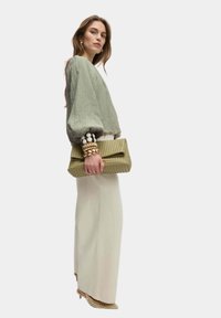 Blusa verde con texture e maniche a sbuffo, abbinata a pantaloni color crema. Tiene in mano una borsa a tracolla intrecciata di un verde chiaro e indossa bracciali impilati.