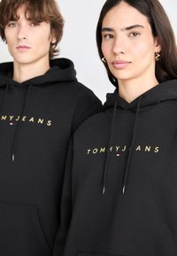 Sudaderas negras con un logo dorado "TOMMYJEANS". Hechas de material suave con bolsillos frontales y cordones. Diseño simple, sin patrones.