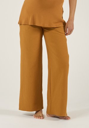 Vrouw die losse, wijd uitlopende mosterdgele broek en bijpassend topje draagt, blootsvoets staand tegen een effen lichte achtergrond.