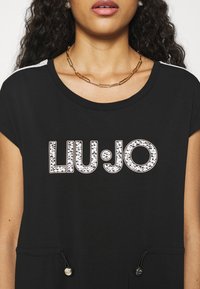 Czarna koszulka z krótkim rękawem z błyszczącym logo "LIU:JO". Zawiera akcenty w postaci sznurków na dolnym brzegu oraz gładką teksturę.