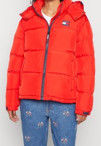 Red puffer jacket with a hood, featuring a front zip closure, quilted texture, and a small brand logo on the chest. Paired with blue jeans.