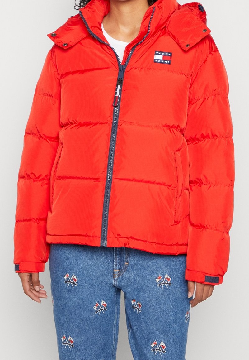 Red puffer jacket with a hood, featuring a front zip closure, quilted texture, and a small brand logo on the chest. Paired with blue jeans.