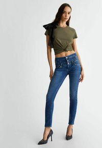 T-shirt cropped verde oliva con un nodo frontale e un volantino in satin nero su una spalla, abbinato a jeans skinny blu e tacchi alti.