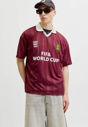 Tânăr purtând un tricou bordo FIFA World Cup, pantaloni scurți din denim deschis la culoare, șapcă neagră, ochelari de soare cu ramă țestoasă și un lanț argintiu pe un fundal simplu.