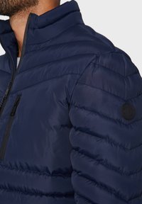 Marineblå dunjakke med quiltet design, høj krave og sort lynlåslomme. Struktureret stof med et cirkulært logo-accents på ærmet.