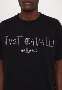 Melnas kokvilnas t-krekls ar baltu, strukturētu uzrakstu "JUST CAVALLI MILANO". Īsas piedurknes un apaļas kakla dizains.