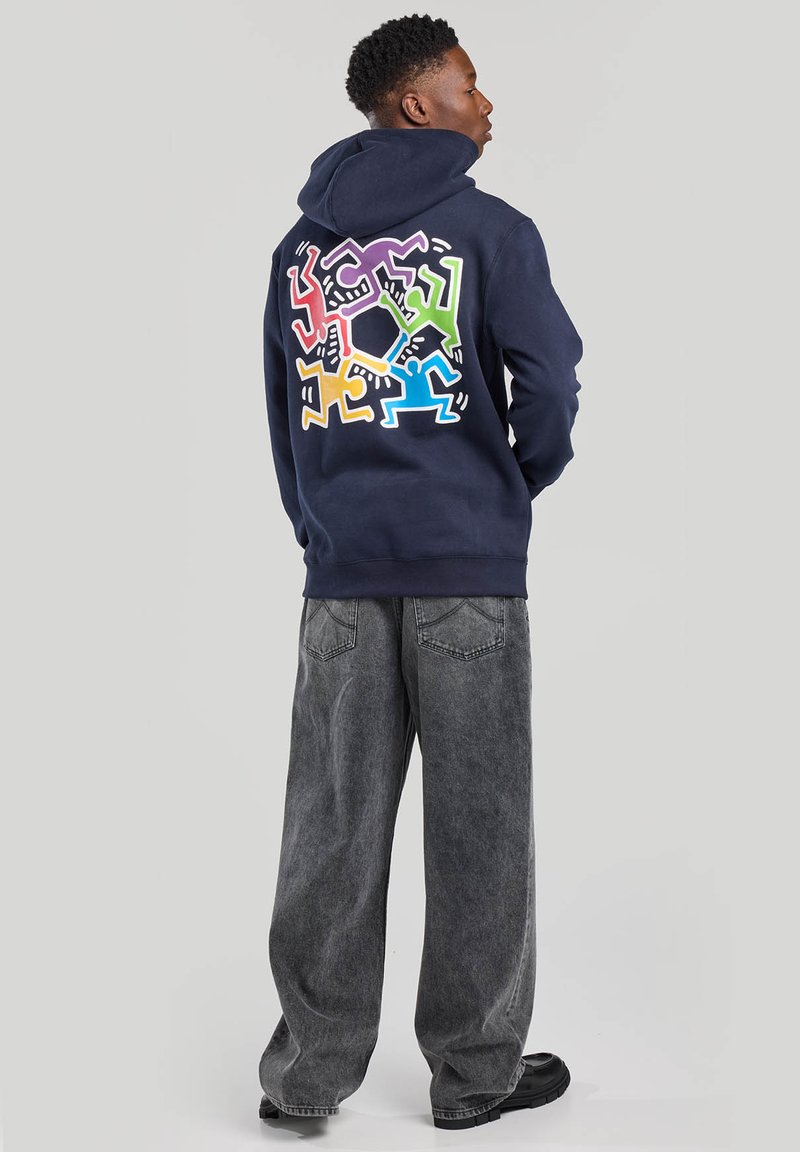 Hoodie bleu marine avec un design graphique coloré au dos, représentant des figures en rose, jaune, vert et bleu sur un fond blanc.