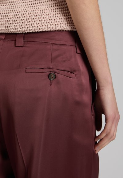 Primer plano de la cadera de una persona que muestra unos pantalones entallados color granate con un bolsillo de ojal abotonado en la parte trasera y una camiseta de punto rosa pálido.