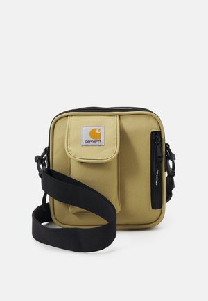 Sac bandoulière compact beige Carhartt avec poche avant à rabat, poche latérale zippée et sangle noire réglable sur fond blanc.