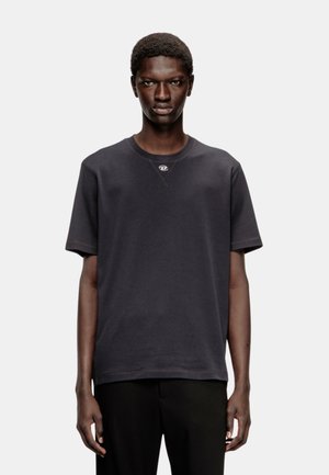 Jeune homme à la peau foncée et aux cheveux courts portant un t-shirt noir uni à manches courtes et un pantalon noir, debout devant un fond clair uni.