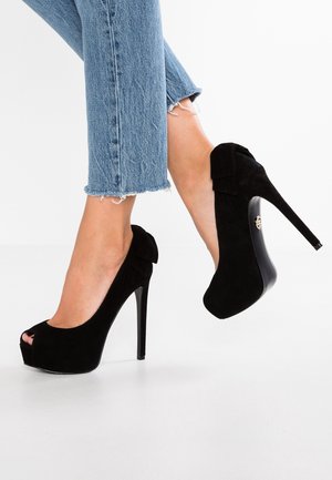 Ben iført korte blå jeans og sorte ruskind peep-toe højhælede sko med platform og sløjfedetalje bagpå.