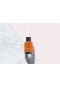 Bottiglia contagocce in vetro trasparente contenente siero arancione, con tappo argento e etichetta con testo bianco, posizionata su uno sfondo di marmo e rosa.